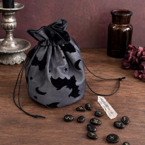 Puede incluir: Una bolsa gris con cordón que presenta un patrón de terciopelo negro con murciélagos, lunas y estrellas. Frente a la bolsa hay diez piedras negras lisas marcadas con símbolos rúnicos dorados y un cristal alargado, transparente y facetado.