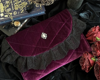 Op de afbeelding: Een dieppaarse fluwelen clutch met een gewatteerd ontwerp en zwarte kanten afwerking. De clutch heeft een decoratieve camee sluiting en staat tegen een donkere, getextureerde achtergrond. Een gouden kroon en een boek zijn ook zichtbaar.