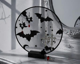 May include: A black and white decorative earring display featuring bat and star designs. The display holds several pairs of earrings with dangling charms. A red gemstone ring sits on the white dresser. A mirror is in the background.