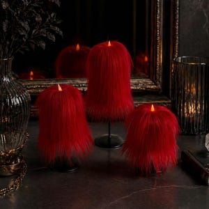 Puede incluir: Tres velas rojas de piel sintética con luces LED parpadeantes. Las velas son de diferentes tamaños y se asientan sobre bases negras. El fondo incluye un espejo, un jarrón y otros elementos decorativos.
