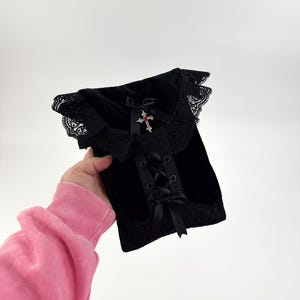 May include: Black velvet corset top with lace trim and a decorative cross. The corset features a lace-up front and satin ribbon bows. The top is a gothic style.