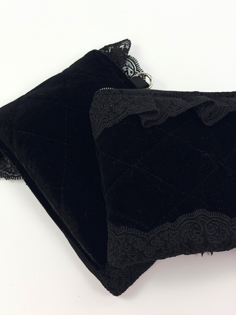 May include: Two black velvet pouches with quilted stitching. The pouches are trimmed with delicate black lace along the top edges. One pouch has a silver ring attached. The pouches are on a white background.