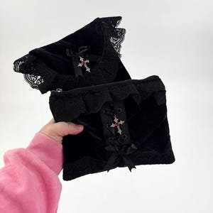 May include: Black velvet clutch with lace trim and a decorative cross pendant with red accents. The clutch features a quilted pattern and a zipper closure. A black bow is attached to the front.