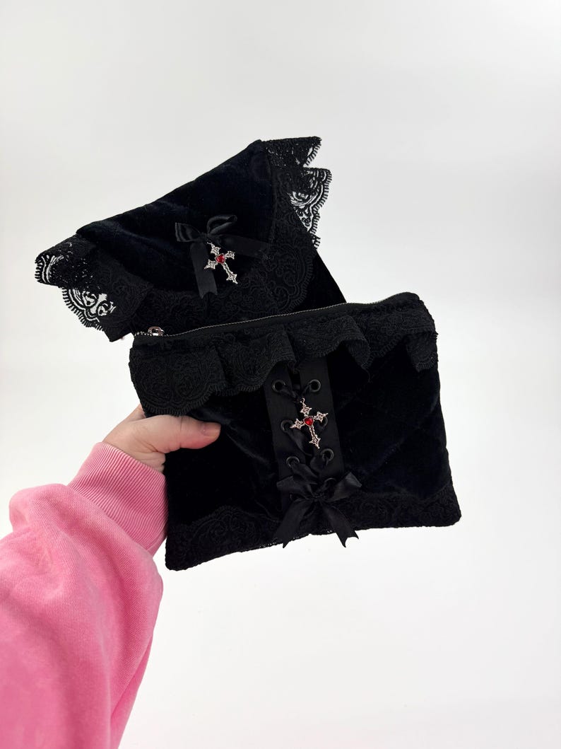 May include: Black velvet clutch with lace trim and a decorative cross pendant with red accents. The clutch features a quilted pattern and a zipper closure. A black bow is attached to the front.