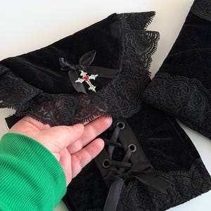 May include: Black velvet arm warmers with lace trim and a decorative cross pendant. The arm warmers feature a lace-up design with black satin ribbon and a small bow. The cross pendant has a red gem in the center.