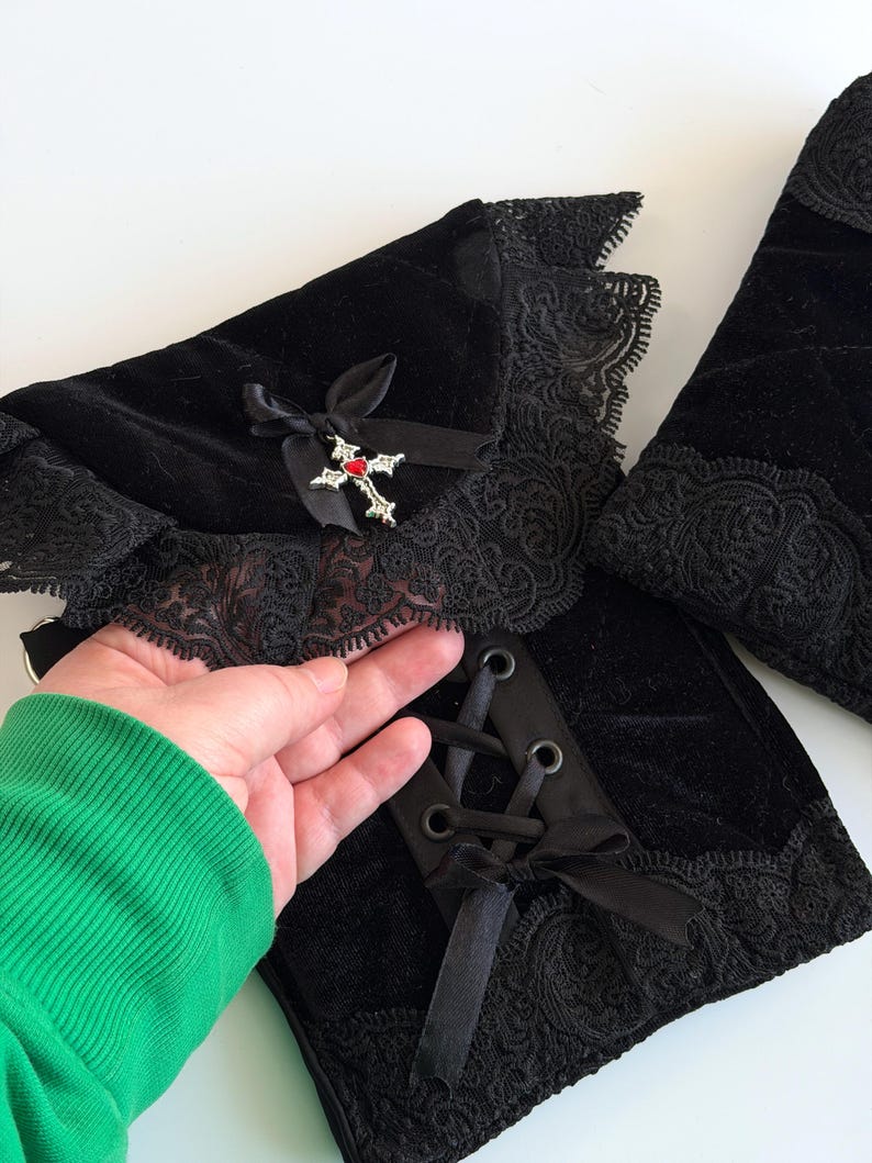 May include: Black velvet arm warmers with lace trim and a decorative cross pendant. The arm warmers feature a lace-up design with black satin ribbon and a small bow. The cross pendant has a red gem in the center.