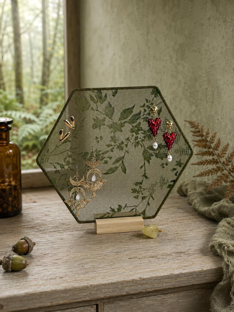 May include: A hexagonal jewelry display stand featuring a green mesh screen with a dark green leaf pattern, supported by a light wood base. The stand holds gold-toned heart studs, red heart-shaped drop earrings with pearls, and gold crescent moon earrings.