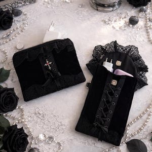 May include: Two black velvet pouches with lace trim and decorative cross accents. One pouch is zippered, the other has a lace-trimmed flap and a corset-style closure. The pouches are displayed with black roses, pearls, and candles.