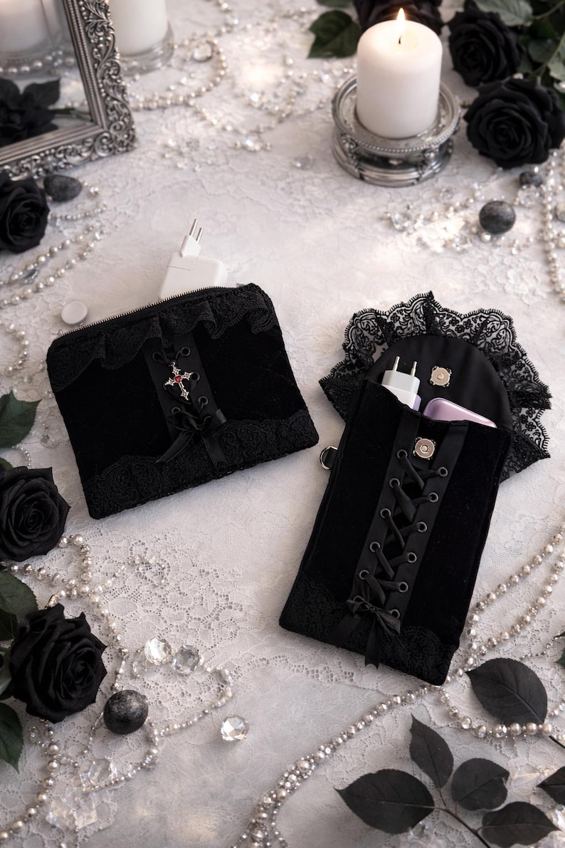 May include: Two black velvet pouches with lace trim and decorative cross accents. One pouch is zippered, the other has a lace-trimmed flap and a corset-style closure. The pouches are displayed with black roses, pearls, and candles.
