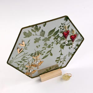 May include: A hexagonal jewelry display stand featuring a green mesh screen with a botanical leaf pattern, supported by a light wood base. The stand holds gold heart earrings, gold moth earrings, and red heart earrings with pearls.