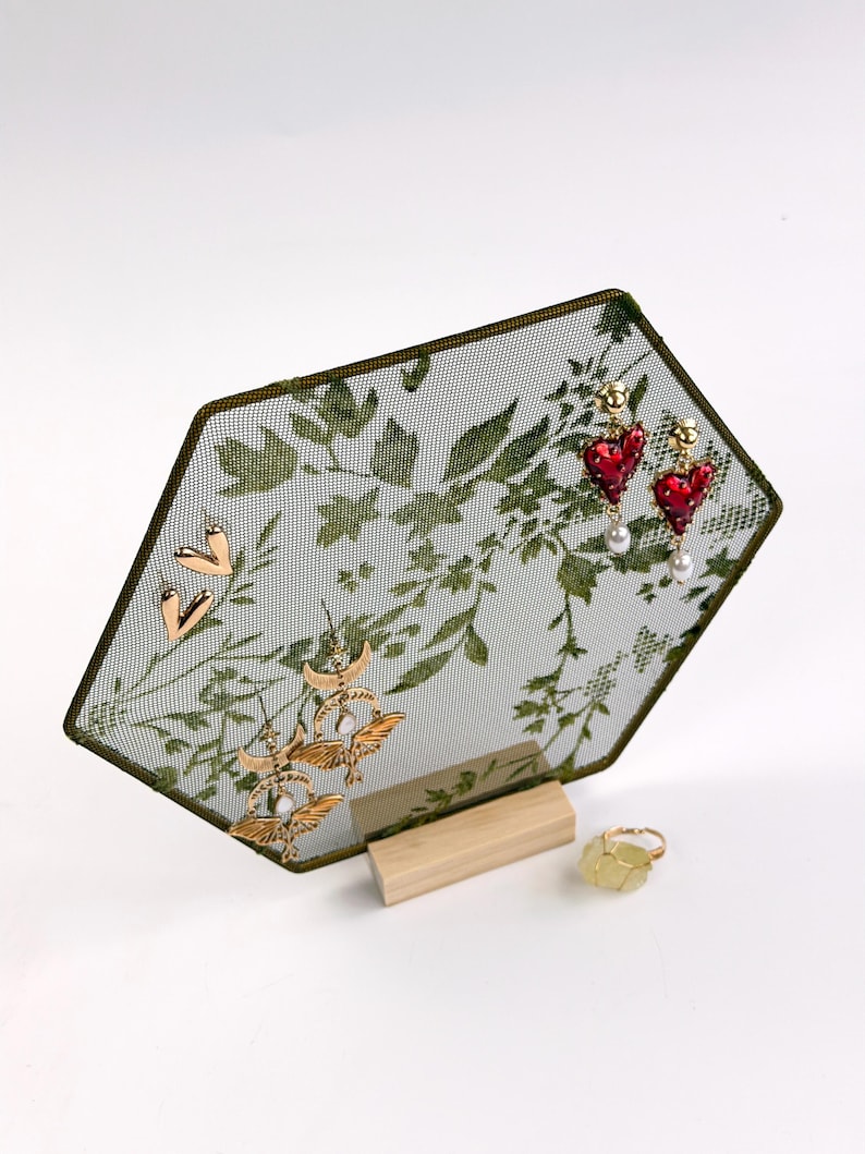 May include: A hexagonal jewelry display stand featuring a green mesh screen with a botanical leaf pattern, supported by a light wood base. The stand holds gold heart earrings, gold moth earrings, and red heart earrings with pearls.