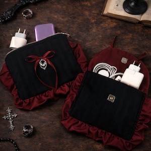 May include: Two black and burgundy pouches with zippers and decorative trim. One pouch has a red ribbon and a silver pendant. The pouches are on a dark wooden surface, along with accessories like a mirror, rings, and a cross.