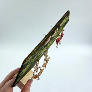 May include: A green and black patterned fabric earring display board held upright by a light wood base. The board features several pairs of dangling earrings, including gold-toned moth designs, red heart-shaped charms, and small white pearl accents.