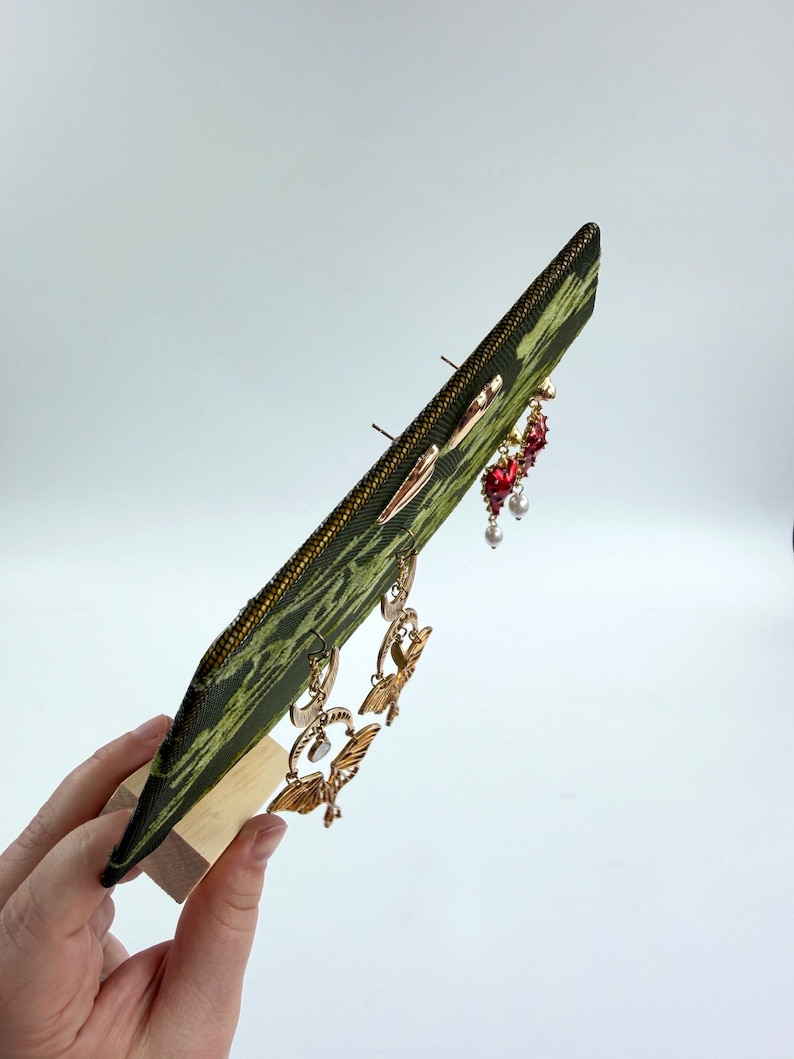 May include: A green and black patterned fabric earring display board held upright by a light wood base. The board features several pairs of dangling earrings, including gold-toned moth designs, red heart-shaped charms, and small white pearl accents.