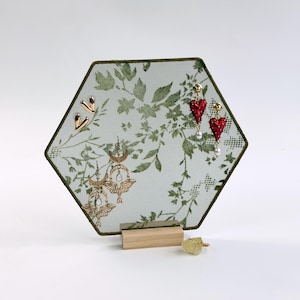 May include: A hexagonal jewelry display stand featuring a green botanical print on a mesh screen with a light wood base. The stand holds two gold-toned heart-shaped studs, two red heart-shaped drop earrings, and two gold-toned moth-shaped earrings.