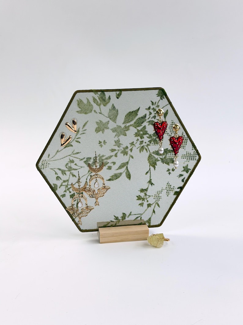 May include: A hexagonal jewelry display stand featuring a green botanical print on a mesh screen with a light wood base. The stand holds two gold-toned heart-shaped studs, two red heart-shaped drop earrings, and two gold-toned moth-shaped earrings.
