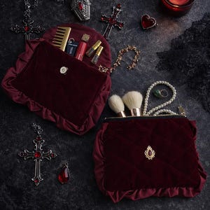 May include: A deep burgundy velvet cosmetic bag with a quilted pattern, open to show makeup brushes and other beauty items. The image also includes gothic jewellery, such as a cross pendant and a heart-shaped ring.