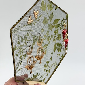 May include: A hexagonal metal mesh jewelry display stand with a green botanical print, featuring gold-tone heart studs, gold-tone moth and moon earrings with white stones, and red heart-shaped earrings with white pearls, set on a wooden base.