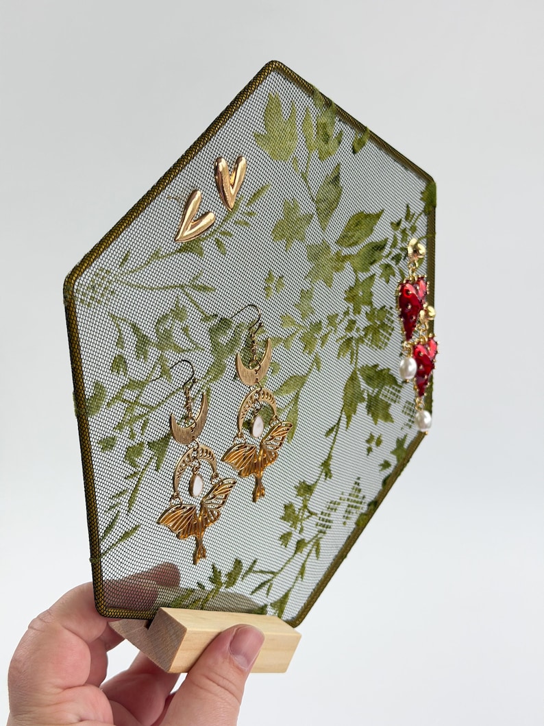 May include: A hexagonal metal mesh jewelry display stand with a green botanical print, featuring gold-tone heart studs, gold-tone moth and moon earrings with white stones, and red heart-shaped earrings with white pearls, set on a wooden base.