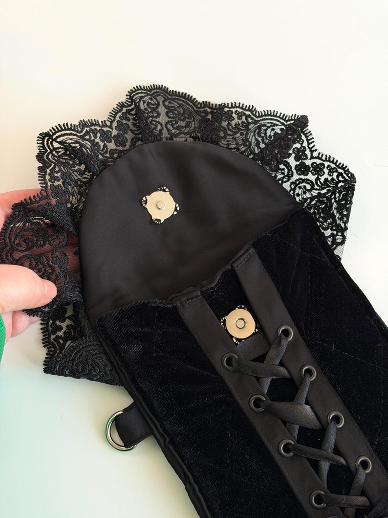 May include: Black velvet corset-style bag with a lace trim. The bag features a satin flap with a magnetic closure and a ribbon lace-up design. A silver-colored metal ring is attached to the side.