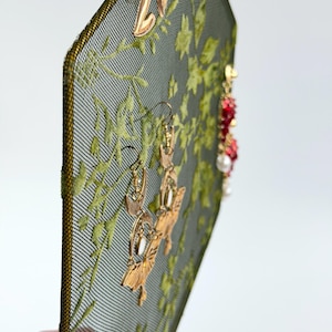 May include: A hexagonal jewelry display stand featuring a dark green mesh screen with an embroidered light green leaf pattern. The stand holds several pairs of earrings, including gold-toned crescent moon shapes and red beaded dangles with white pearls.