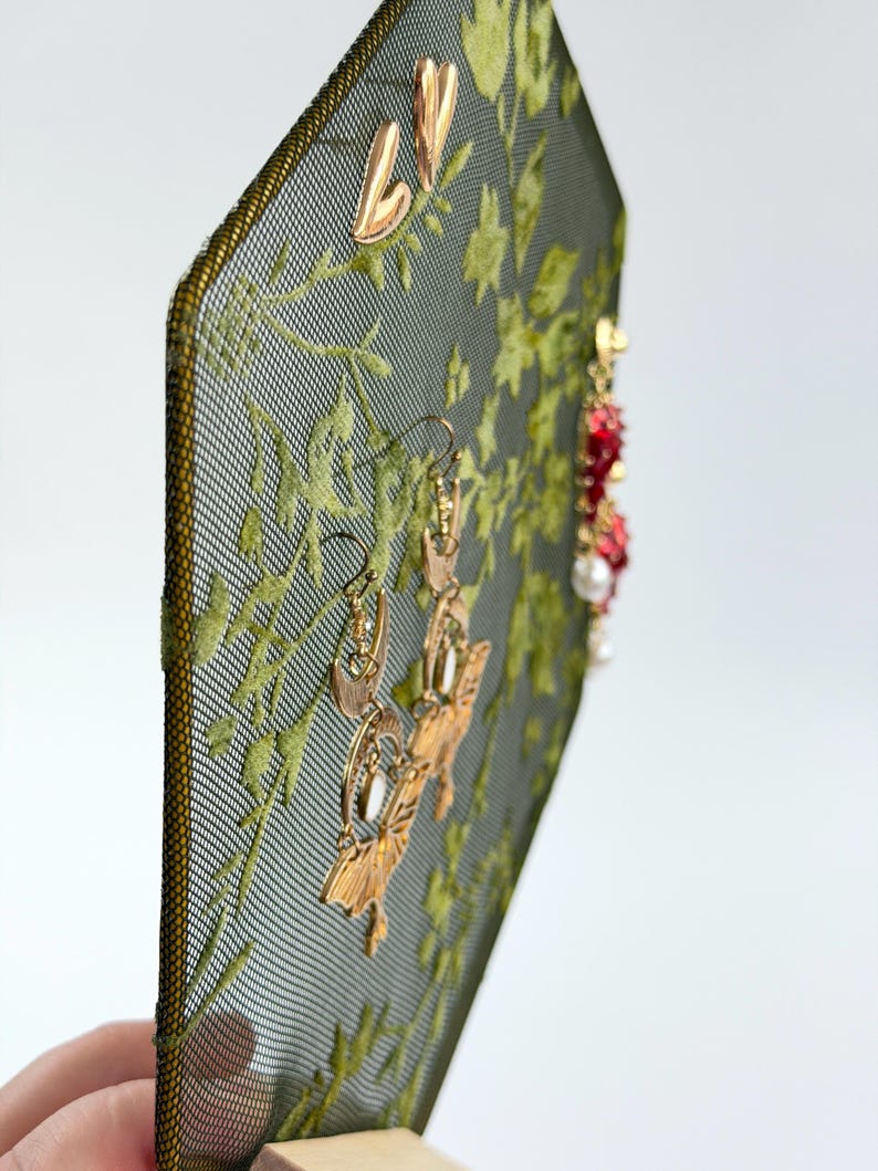 May include: A hexagonal jewelry display stand featuring a dark green mesh screen with an embroidered light green leaf pattern. The stand holds several pairs of earrings, including gold-toned crescent moon shapes and red beaded dangles with white pearls.