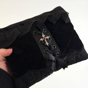 May include: Black velvet accessory with a lace trim. The center features a black ribbon with a decorative cross and red gem. The accessory has a quilted pattern and is tied with black satin ribbons.