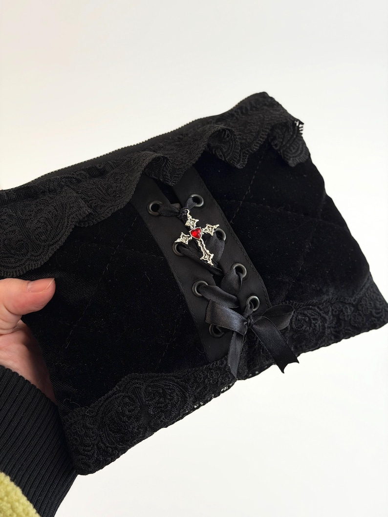May include: Black velvet accessory with a lace trim. The center features a black ribbon with a decorative cross and red gem. The accessory has a quilted pattern and is tied with black satin ribbons.