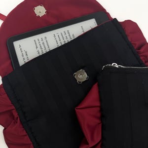 May include: A burgundy satin clutch with a black, vertically striped interior. The clutch is open, revealing an e-reader with text. A small black zippered pouch is visible. The clutch has a ruffled edge and a magnetic clasp.