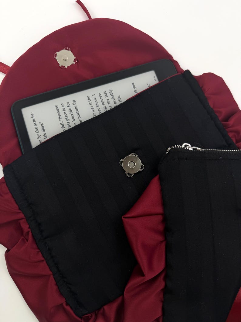 May include: A burgundy satin clutch with a black, vertically striped interior. The clutch is open, revealing an e-reader with text. A small black zippered pouch is visible. The clutch has a ruffled edge and a magnetic clasp.