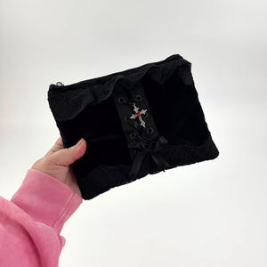 May include: Black velvet clutch with lace trim and a decorative cross pendant. The clutch features a corset-style ribbon detail and a small bow. The bag is rectangular and has a zipper closure.
