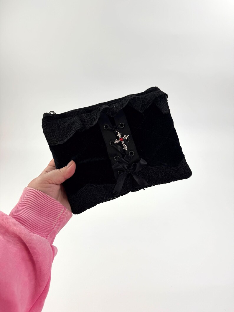 May include: Black velvet clutch with lace trim and a decorative cross pendant. The clutch features a corset-style ribbon detail and a small bow. The bag is rectangular and has a zipper closure.