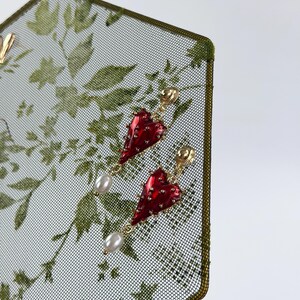 May include: A pair of gold-tone earrings featuring a glossy red heart-shaped charm with small metallic studs and a dangling white teardrop pearl. The earrings are secured with a gold-tone post back and are displayed on a green mesh surface.