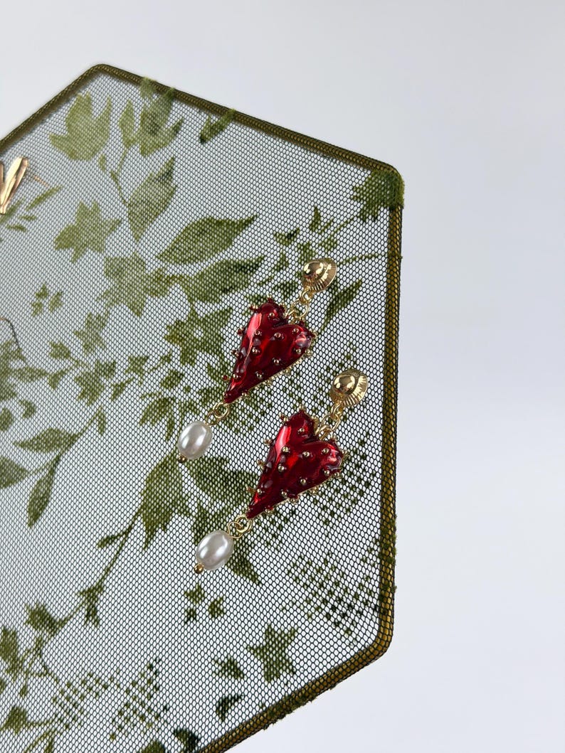 May include: A pair of gold-tone earrings featuring a glossy red heart-shaped charm with small metallic studs and a dangling white teardrop pearl. The earrings are secured with a gold-tone post back and are displayed on a green mesh surface.