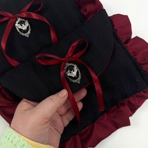 May include: Black fabric pouch with a red satin ribbon and a silver bat-shaped brooch. The pouch has a ruffled burgundy trim. The design is gothic-inspired and features a striped pattern on the black fabric.