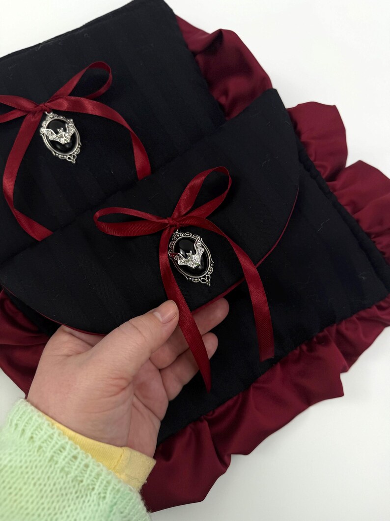 May include: Black fabric pouch with a red satin ribbon and a silver bat-shaped brooch. The pouch has a ruffled burgundy trim. The design is gothic-inspired and features a striped pattern on the black fabric.