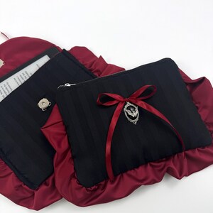 May include: Two black rectangular pouches with vertical stripes, trimmed with ruffled burgundy fabric. One pouch has a silver zipper and a burgundy ribbon with a decorative charm. The other pouch has a silver button.