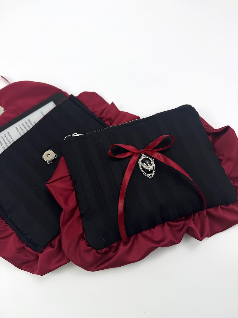 May include: Two black rectangular pouches with vertical stripes, trimmed with ruffled burgundy fabric. One pouch has a silver zipper and a burgundy ribbon with a decorative charm. The other pouch has a silver button.