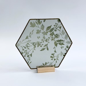 May include: A hexagonal metal mesh screen featuring a green botanical print of leaves and flowers. The screen is framed in a thin dark metal border and is supported by a rectangular light-colored wooden base that holds it in an upright position.