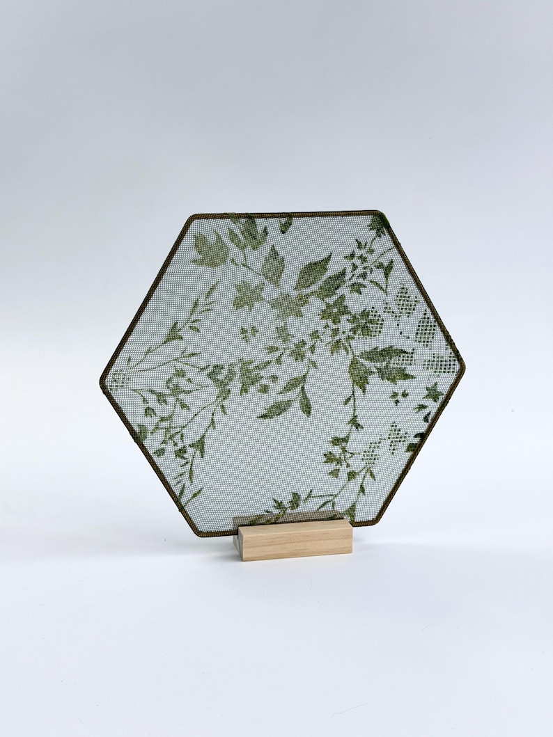 May include: A hexagonal metal mesh screen featuring a green botanical print of leaves and flowers. The screen is framed in a thin dark metal border and is supported by a rectangular light-colored wooden base that holds it in an upright position.