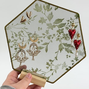 May include: A hexagonal earring display stand with a green leaf pattern on a mesh screen, featuring gold-tone metal heart studs, moth-shaped drop earrings with crescent moons and white stones, and red heart-shaped drop earrings with white pearls.