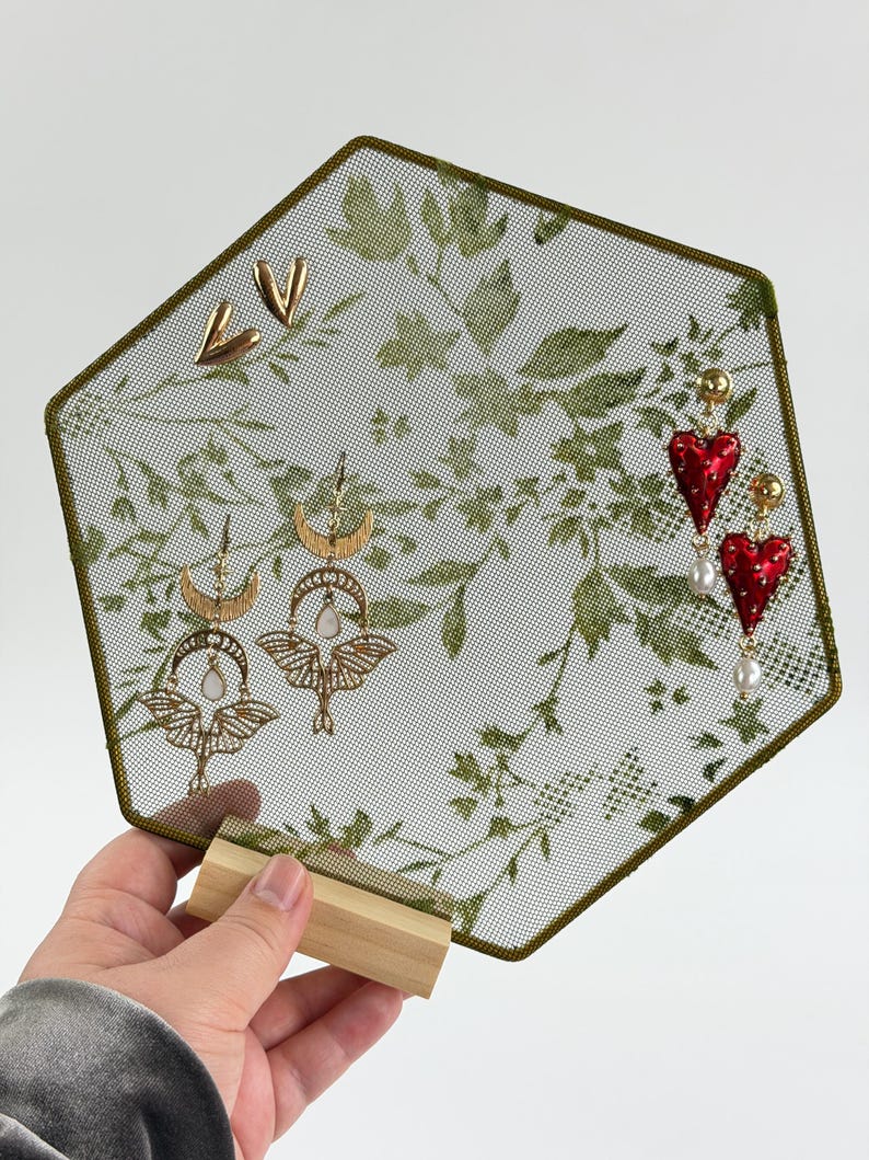 May include: A hexagonal earring display stand with a green leaf pattern on a mesh screen, featuring gold-tone metal heart studs, moth-shaped drop earrings with crescent moons and white stones, and red heart-shaped drop earrings with white pearls.