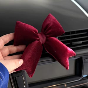 May include: A burgundy velvet bow, likely for decoration, is held against a black car dashboard. The bow has a classic shape with multiple loops and tails, and appears to be made of a soft, plush material.
