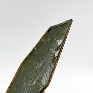 May include: A rectangular earring display stand featuring a dark green mesh screen with a gold-toned metal frame. The mesh surface holds multiple pairs of earrings, secured with clear plastic earring backs visible on the front side of the display.