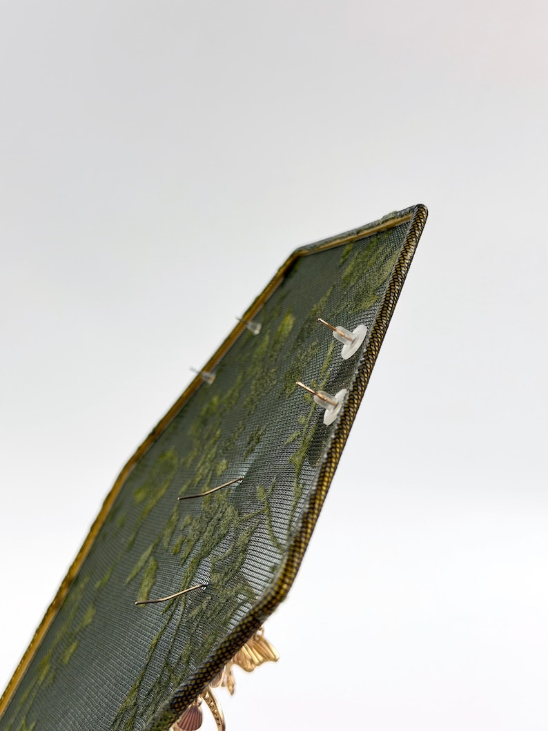 May include: A rectangular earring display stand featuring a dark green mesh screen with a gold-toned metal frame. The mesh surface holds multiple pairs of earrings, secured with clear plastic earring backs visible on the front side of the display.