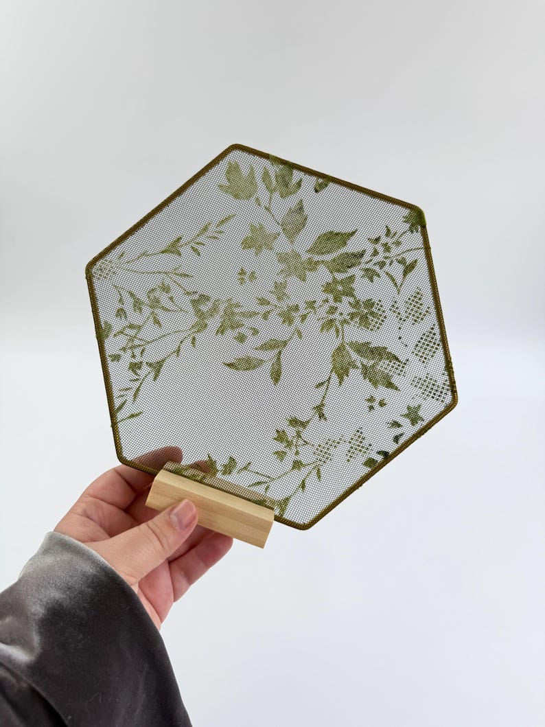 May include: A hexagonal mesh display panel featuring a green botanical leaf and floral pattern. The mesh is framed in a thin green border and is supported by a small, rectangular light-colored wooden base stand for upright positioning.
