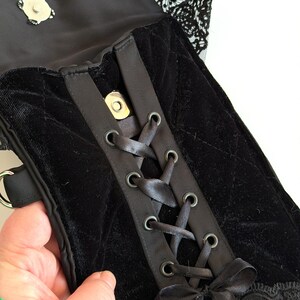 May include: Black velvet handbag with a corset-style front, featuring satin ribbon lacing and metal eyelets. The bag has a quilted pattern and is trimmed with black lace. A silver clasp secures the top flap.