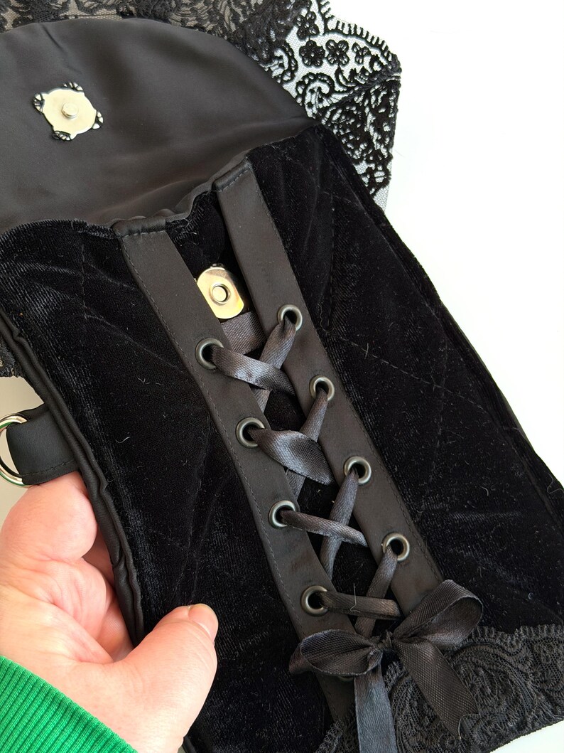 May include: Black velvet handbag with a corset-style front, featuring satin ribbon lacing and metal eyelets. The bag has a quilted pattern and is trimmed with black lace. A silver clasp secures the top flap.