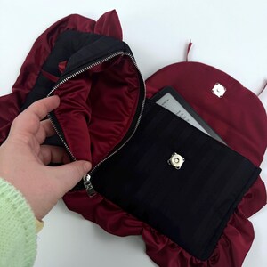 May include: Black and burgundy clutch bag with a zipper closure, and a matching burgundy satin lining. The bag is open, revealing a black case with an e-reader inside. The bag has a unique shape with ruffled edges.