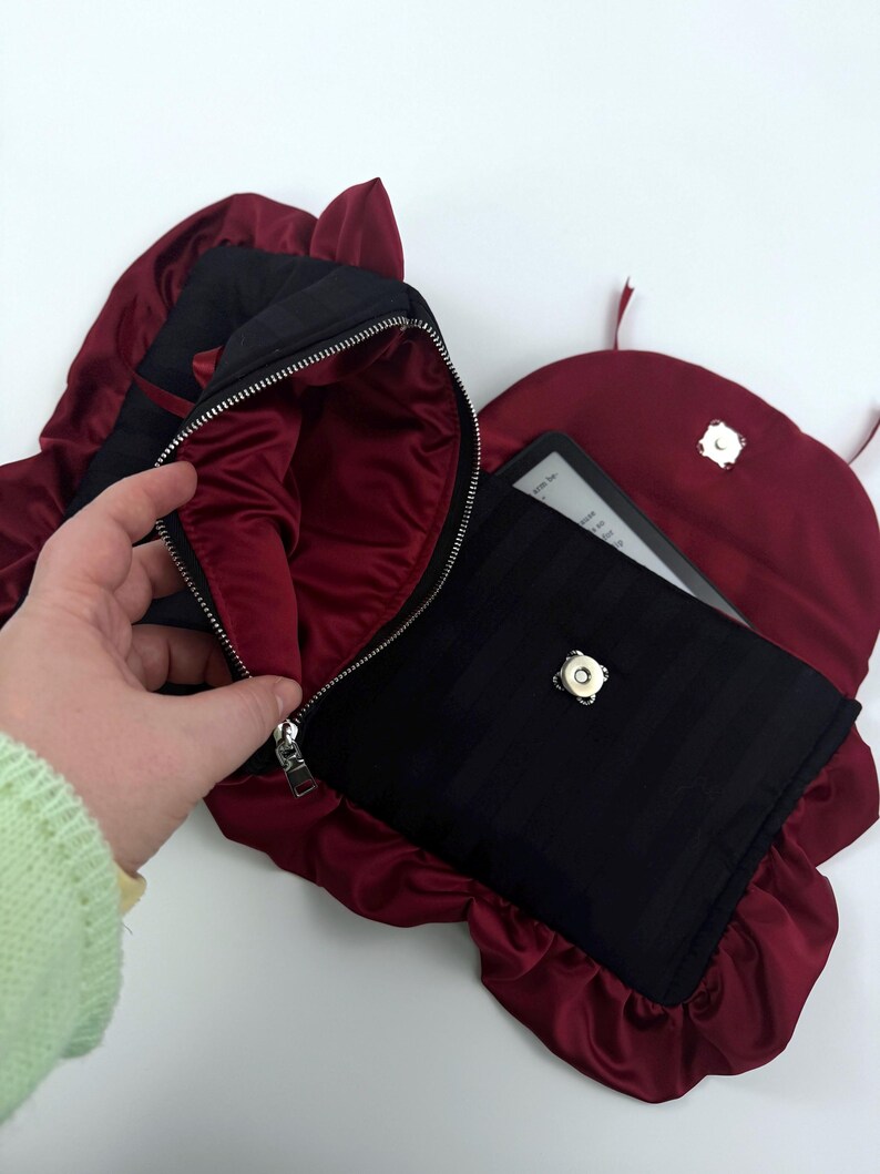 May include: Black and burgundy clutch bag with a zipper closure, and a matching burgundy satin lining. The bag is open, revealing a black case with an e-reader inside. The bag has a unique shape with ruffled edges.